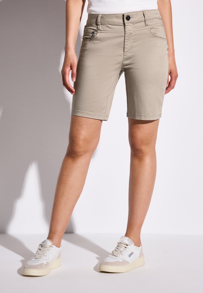 Shorts beige à coupe slim, avec poches avant et un petit détail de fermeture éclair sur le côté. Fabriqués en tissu léger, associés à des baskets blanches.