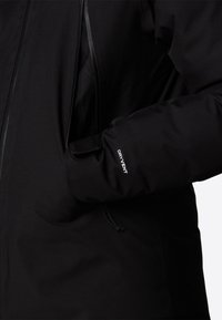 Chaqueta negra impermeable con cremallera en la parte delantera, un gran bolsillo y un logo "DRY VENT" en la manga. Textura suave y mate.