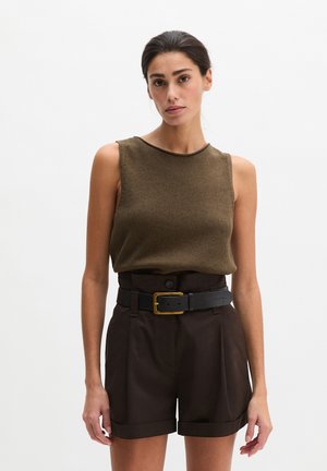 Vrouw draagt een mouwloos bruin gebreid topje, gestopt in een hoogwater donkerbruine korte broek met een zwarte riem met een gouden gesp.