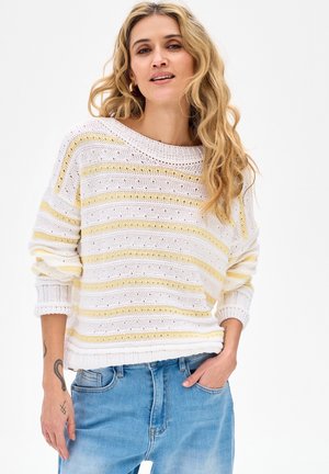 Kobieta z długimi, falowanymi blond włosami, ubrana w biało-żółty sweter w paski i niebieskie dżinsy, z jedną ręką w kieszeni.