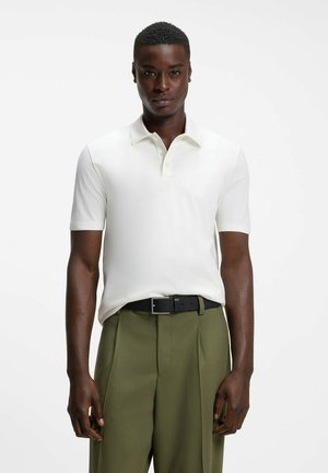Uomo con capelli corti indossa una polo bianca a maniche corte infilata in pantaloni verde oliva con cintura nera, in piedi su sfondo bianco.