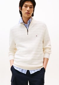 Cremefarbenes gestricktes Sweatshirt mit horizontalen Streifen, ausgestattet mit einem Reißverschlusskragen und gerippten Bündchen, getragen über einem blau-weißen gestreiften Hemd.