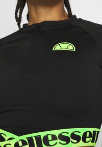 Svart träningströja med halvnäckslås, strukturerad tyg och en neongrön kant med kontrasterande svart text. Logotyp på bröstet.