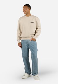Beige textured sweatshirt with black logo, paired with light blue baggy jeans and white sneakers, posing on a white background.