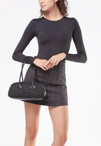 Borsa in suede nera con dettagli in pelle liscia, forma arrotondata e due manici. Il modello indossa un top nero aderente e una mini gonna a quadri.
