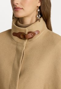 Καμηλοπόδαρο παλτό με ψηλό γιακά, με καφέ δερμάτινη λεπτομέρεια και χρυσό κομβίο. Υφασμάτινη υφή με λεία επιφάνεια.