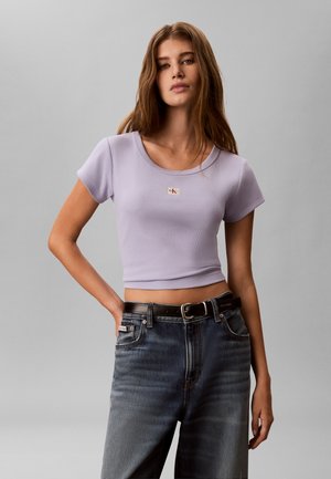 Joven mujer con cabello largo y castaño lleva un crop top ajustado color lavanda y vaqueros lavados oscuros con cinturón negro, posando de forma neutral sobre un fondo gris.
