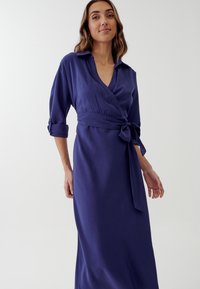 Abito avvolgente blu navy realizzato in tessuto morbido, con collo, maniche lunghe arrotolate ai polsini e vita accentuata da un nastro.