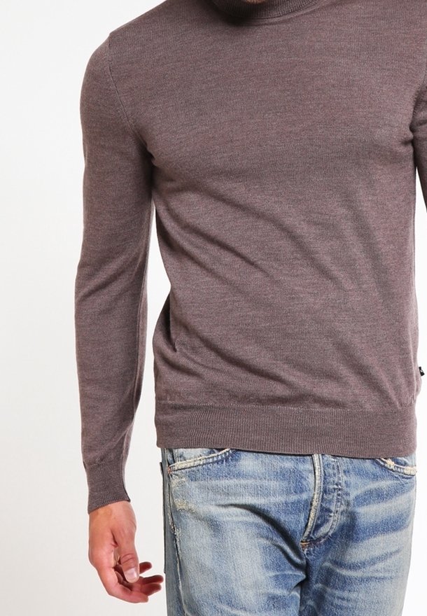Homme portant un pull marron moulant à manches longues et un jean bleu clair délavé, avec son bras gauche détendu le long du corps.