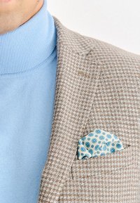 Brązowa marynarka w wzór houndstooth z jasnoniebieskim golfem underneath. Chusteczka w kieszeni ma niebiesko-kremowy wzór geometryczny.