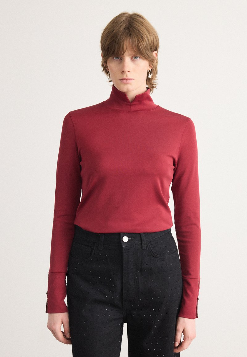 Red long-sleeve high-neck top, fitted design, smooth fabric. Paired with high-waisted black jeans featuring dotted accents.
