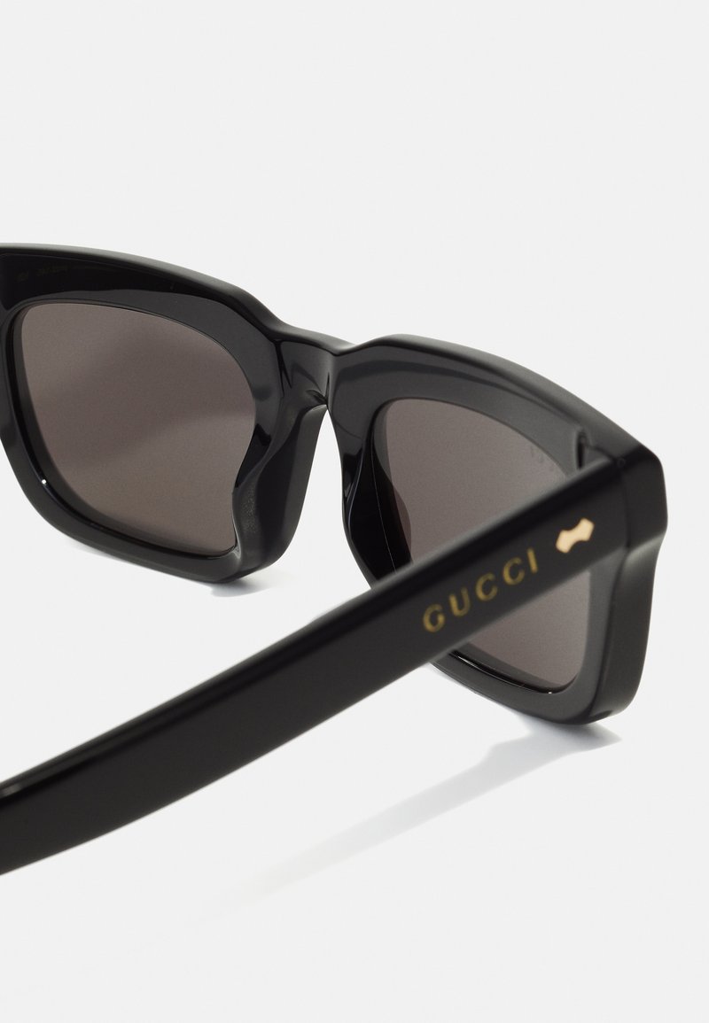 Occhiali da sole neri oversize con montatura angolare e lenti scure. L'asta presenta un logo "GUCCI" dorato e un delicato dettaglio decorativo.