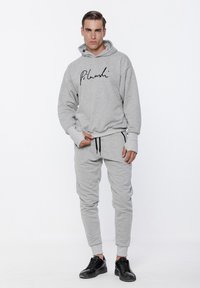 Szara bluza z kapturem z czarnym logo w stylu pisma, zestawiona z pasującymi dresami. Posiada ściągacz w talii oraz prążkowane mankiety. Włożona z czarnymi trampkami.