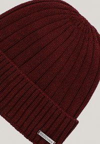 Bordowa dziana czapka z prążkowanym wzorem i złożonym mankietem. Zawiera metalową naszywkę z logo przeszytą z boku.