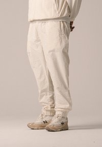 Jogging beige avec des poignets élastiques, un tissu texturé et des poches, associé à des baskets New Balance beiges avec un logo noir.