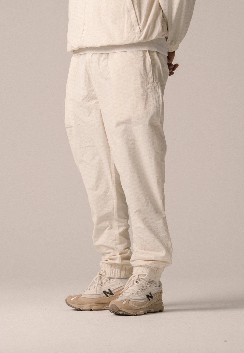 Jogging beige avec des poignets élastiques, un tissu texturé et des poches, associé à des baskets New Balance beiges avec un logo noir.