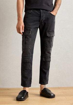 Pantalones cargo negros de mezclilla con múltiples bolsillos de parche, diseño de pierna recta y ligeras marcas de desgaste. Combinados con zuecos negros.