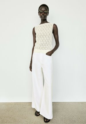 Donna con top in maglia crema senza maniche, pantaloni bianchi larghi, sandali neri e occhiali da sole scuri, in piedi davanti a uno sfondo bianco semplice.