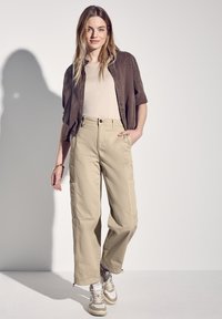 Beige cargo kalhoty s volným střihem; zkombinované se světle béžovým topem a tmavohnědou košilí s krátkým rukávem na knoflíky. Bílé a béžové tenisky.