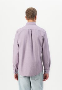 Mann, der mit dem Rücken zur Kamera steht, trägt ein helllila Langarmhemd und hellblaue Jeans vor einem schlicht weißen Hintergrund.