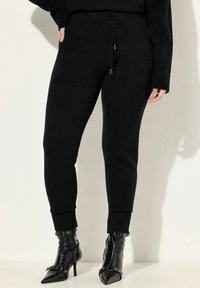 Černé pletené joggery s opaskem na šňůrku, žebrovanými manžetami a hladkým povrchem, spárované s elegantními černými špičatými kotníkovými botami s kovovými akcenty.