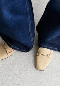 Mocassini beige in pelle scamosciata con un'accento in catena dorata, abbinati a jeans scuri a gamba larga, su una superficie testurizzata grigia chiara.