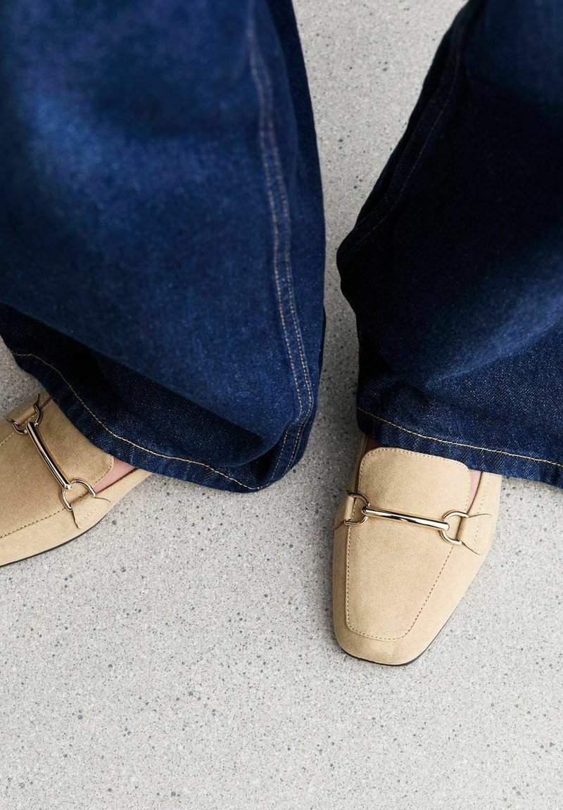 Mocassini beige in pelle scamosciata con un'accento in catena dorata, abbinati a jeans scuri a gamba larga, su una superficie testurizzata grigia chiara.