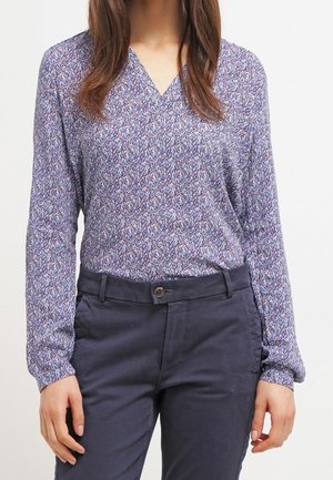 Femme portant un chemisier bleu à motifs, manches longues, col en V, rentré dans un pantalon bleu marine foncé avec un bouton et des passants pour ceinture.