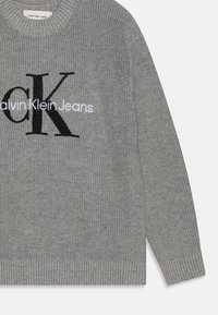 Maglione grigio lavorato a maglia con texture a coste, caratterizzato da un grande logo 'CK' nero e la scritta 'Calvin Klein Jeans' in bianco. Design classico a girocollo.