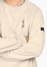 Felpa beige a maniche lunghe con tasca sul petto con zip, maniche raglan e tessuto testurizzato. Presenta una patch con logo sulla manica sinistra.