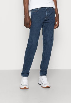 Jeans in denim blu scuro con vestibilità dritta, caratterizzati da un design a cinque tasche e cuciture a contrasto, abbinati a sneakers bianche.