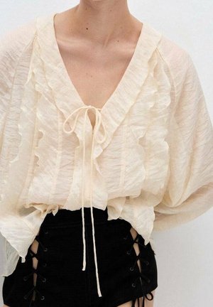 Woman wearing a sheer cream ruffled blouse with tie front, paired with high-waisted black shorts featuring side lace-up details.