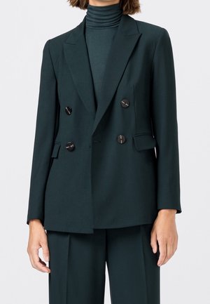 Blazer croisé vert foncé avec boutons noirs, porté sur un haut à col montant assorti et un pantalon à jambes larges.