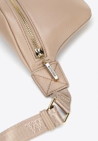 Beżowa skórzana torba na ramię z teksturowaną powierzchnią, złotym zamkiem błyskawicznym i odpinanym materiałowym paskiem z akcentem logo na okuciach.