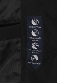 Zwart stoffen label met pictogrammen voor winddicht, waterdicht (5000 mm), ademend (3000 mvp) en volledig afgeplakte naden. Tekst is wit op een donkere achtergrond.