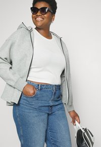 Smaidīga sieviete, kas valkā saulesbrilles, baltu tvīkdāmu, pelēku zipsvārku un zilo džinsu, ar vienu roku tur melnbaltu roku somu.