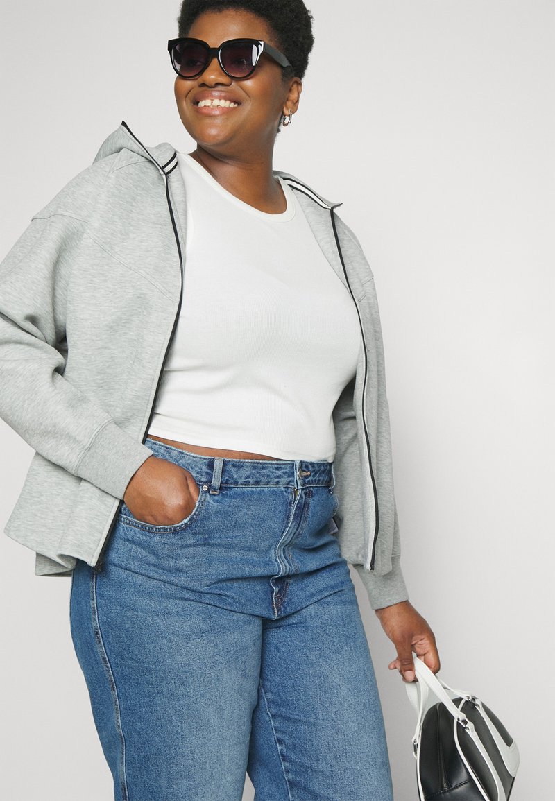 Femme souriante portant des lunettes de soleil, un débardeur blanc, un sweat à capuche gris zippé et un jean bleu, tenant un sac à main noir et blanc d'une main.
