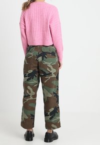 Personne portant un pull rose côtelé et un pantalon ample à motif camouflage avec de grandes poches arrière, debout devant un fond blanc uni.