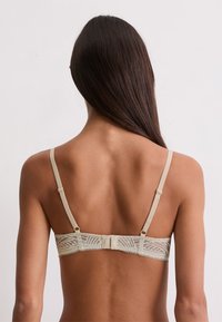 Donna con lunghi capelli scuri vista da dietro, indossa un reggiseno in pizzo beige con spalline regolabili su uno sfondo neutro.