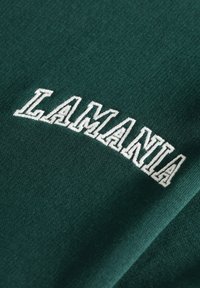 Žali audinys su baltais siuvinėtais „LAMANIA“ užrašais, turintis lygią tekstūrą ir šiek tiek išlenktą formą. Dizainas yra paprastas ir aiškus.