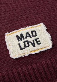 Burgunder strikket stof med en tekstureret cremefarvet lap, der har "MAD LOVE" i fed sort skrift med gul kantstikning. Kanterne er flossede.