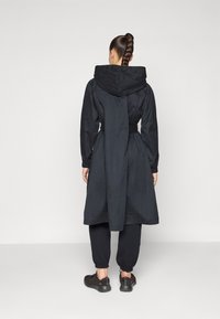 Black hooded coat with a cinched waist, knee-length cut, and textured fabric. Paired with black joggers and dark shoes. Visible back detail.