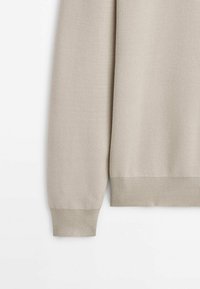 Manche de pull en maille beige et ourlet inférieur avec poignets et ceinture côtelés sur fond blanc.