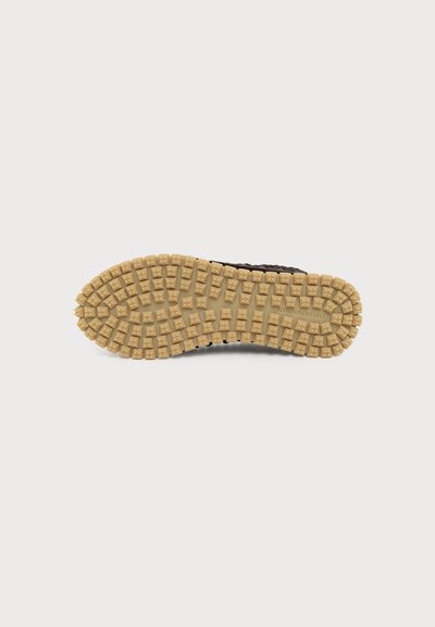Semelle de chaussure avec une épaisse bande de roulement en caoutchouc beige présentant des crampons carrés sur une chaussure marron, montrée sur un fond blanc.