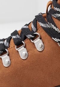 Botte en daim marron avec des lacets rayés noirs et blancs passés à travers des œillets argentés, présentant une surface texturée et des coutures détaillées.