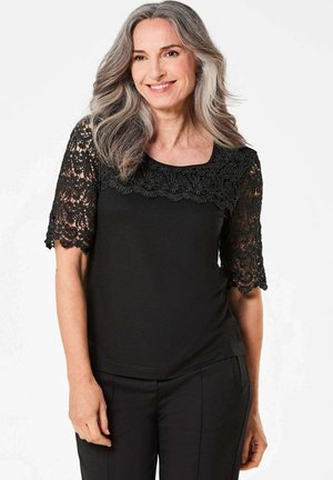 Schwarzes Oberteil mit kurzen Spitzenärmeln, das ein Blumenmuster auf der Spitze aufweist und über einen runden Halsausschnitt verfügt, kombiniert mit schwarzen Hosen.