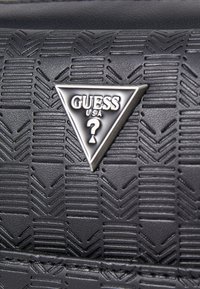 Material negro texturizado con un patrón geométrico, con un logo triangular plateado "GUESS USA" incrustado en el centro.