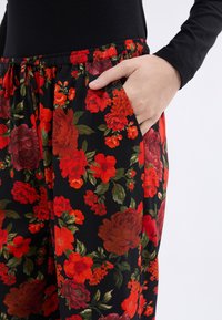 Pantalones florales con un fondo negro y grandes flores rojas y naranjas, detalle de bolsillo y una cintura elástica con cordón.