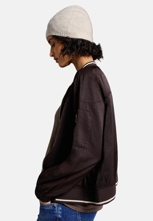 Donkerbruine bomberjack met elastische manchetten, witte accenten en een rits. Gecombineerd met een lichtbeige gebreide beanie en een grijze top.
