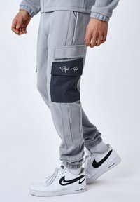 Pantaloni della tuta grigi con tasche cargo, pannelli a contrasto blu navy, polsini a coste e dettaglio del logo sulla tasca; abbinati a sneakers bianche Nike.